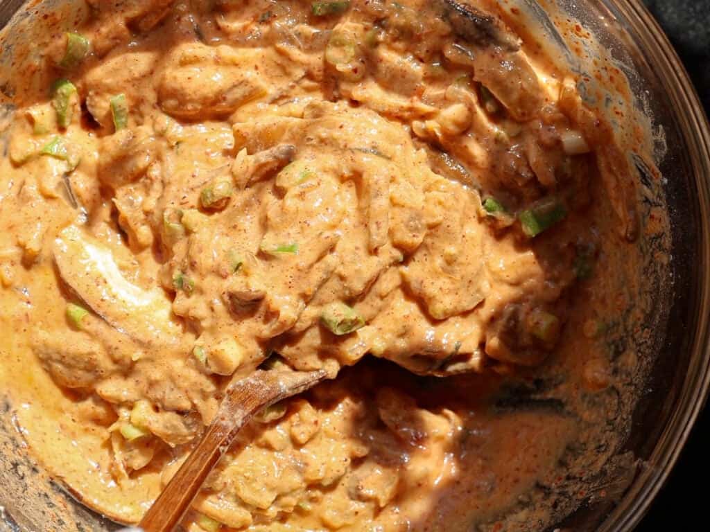 A close-up of a bowl filled with a thick, orange-tinted mixture containing chopped green herbs or vegetables, with a wooden spoon partially submerged in the mixture.